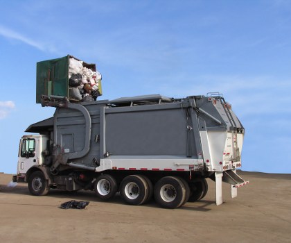 A large industrial waste collection truck parked on an open, paved surface with a clear blue sky in the background. The truck has a white cab with a black bumper and side mirrors, and an extended grey waste storage container mounted on the chassis. On top of the container, there is a green compactor unit with white and black waste materials securely compressed inside, visible through the open flap at the rear. The machinery appears designed for commercial rubbish collection, with multiple black wheels supporting the heavy-duty vehicle. Nearby on the ground, a small pile of loose rubbish, likely discarded waste materials, is visible. The setting suggests a waste management operation, with the truck ready for rubbish removal service, consistent with services offered by Commercial Waste Wood Green. The environment is an outdoor industrial or storage yard area with no other structures or vehicles immediately visible, emphasizing the truck's role in commercial waste collection in the nearby area of Wood Green, London.