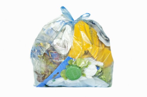A transparent plastic rubbish bag filled with a mix of waste materials, including yellow packaging, a green plastic bottle, white and green plastic items, and paper or cardboard pieces. The bag is placed on a surface with a plain white background, showing the varied textures and colours of the waste. The packaging appears crinkled and tightly packed, with some items visibly bulging through the bag's semi-transparent film. The waste suggests general household refuse that could be collected during commercial waste removal services in the Wood Green area. The image reflects typical rubbish collected by companies like Commercial Waste Wood Green as part of their general rubbish removal operations. There are no additional objects or environmental context visible beyond the bag itself, emphasizing the contents' diversity and the importance of proper waste disposal in urban environments.
