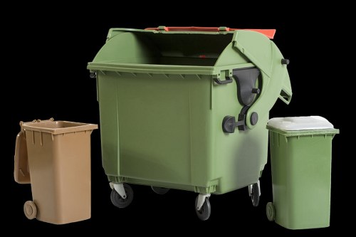 A large green wheeled commercial rubbish bin with a closed lid, positioned on a dark background, accompanied by two smaller closed bins—one beige and one light green—placed on either side. The green bin appears to be made of durable plastic with a smooth finish and features a black handle on the front, while the smaller bins also have lids and are situated outdoors on a paved or concrete surface. The environment suggests a waste collection area, relevant to services like those offered by Commercial Waste Wood Green for rubbish removal in the local area. The overall composition emphasizes waste management containers used for general refuse collection, reflecting typical equipment used by businesses or residential properties in the London borough of Haringey, which includes Wood Green postcode areas.