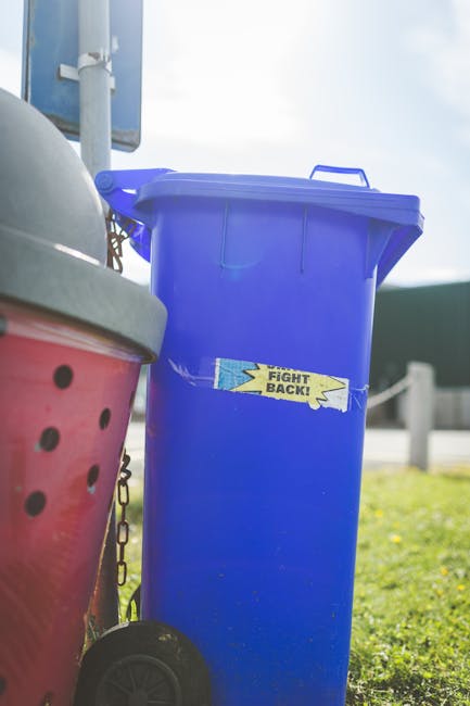 A row of large plastic wheelie bins lined up along a pavement adjacent to a red-brick wall, with each bin featuring a removable lid. The bins are predominantly blue, with some green and red-colored li